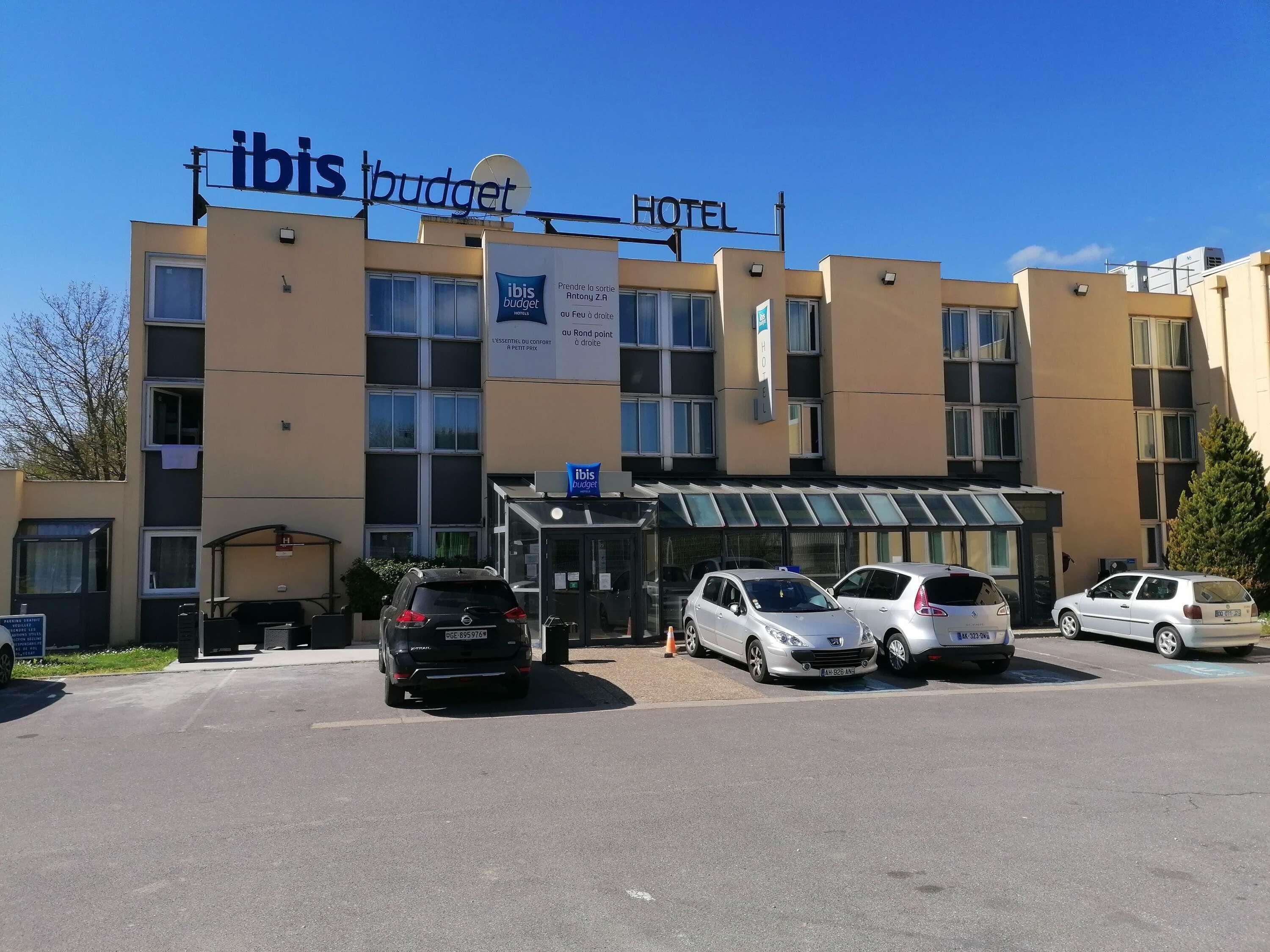 Un hotel con un cartel que dice 'ibis budget HOTEL' y un banner que anuncia 'ibis budget'. Hay varios coches estacionados en la parte delantera.