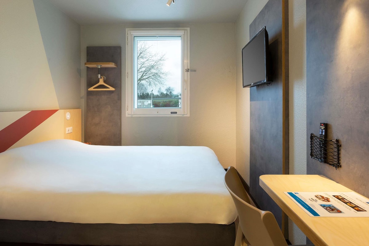 A hotel room with a bed, a desk, a TV, a window, and a hanging coat rack.
