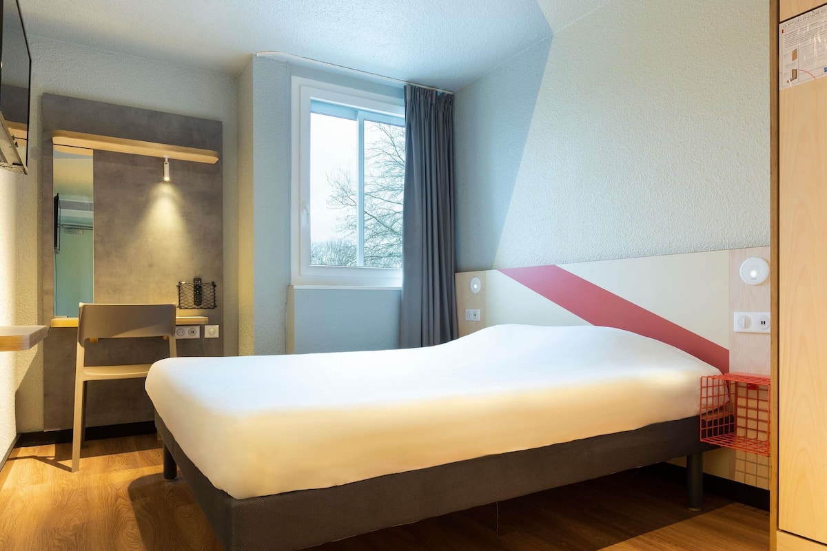 A hotel room with a bed, a desk, a chair, a window with curtains, and a red basket.