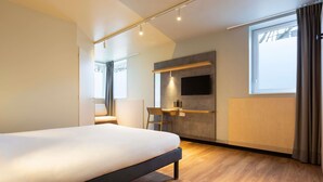 A modern hotel room with a large bed, a desk, a chair, a TV mounted on the wall, and a window with curtains. - ibis Budget Antony Massy (renovated 2024) (Antony)