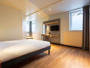 A modern hotel room with a large bed, a desk, a chair, a TV mounted on the wall, and a window with curtains. - ibis Budget Antony Massy (renovated 2024) (Antony)