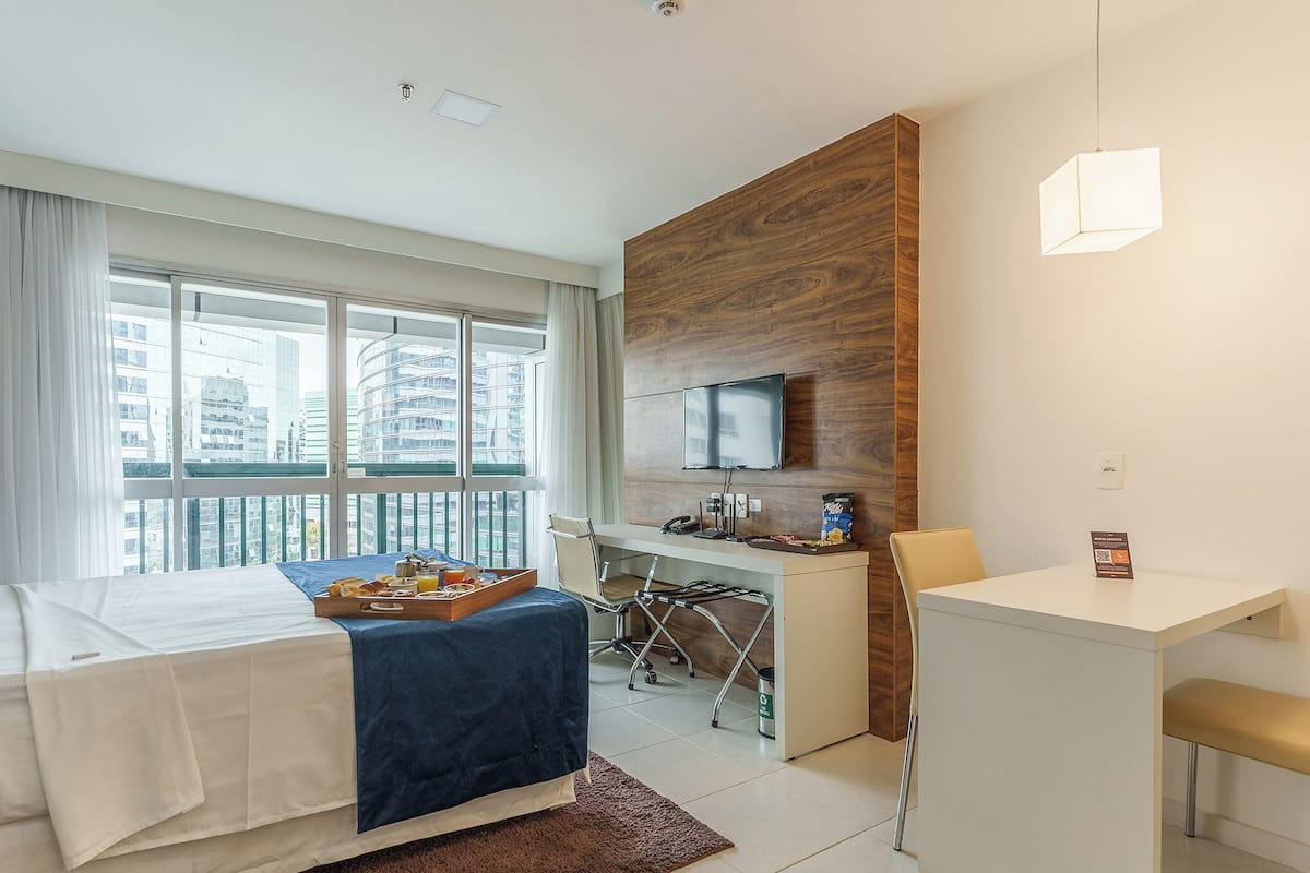 A hotel room with a bed, a desk, a chair, a TV, and a window with a city view.