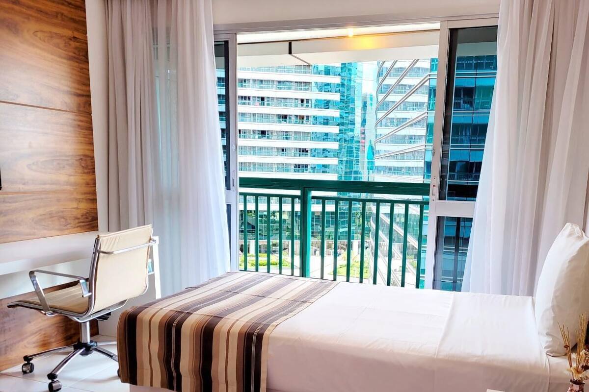 A hotel room with a bed, a desk, a chair, and a balcony with a view of high-rise buildings.