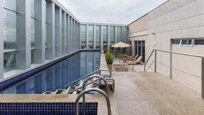 A rooftop pool area with a clear view of the city, a seating area with chairs and an umbrella, and a pool with blue tiles. - Hplus Vision Executive (Brasília)