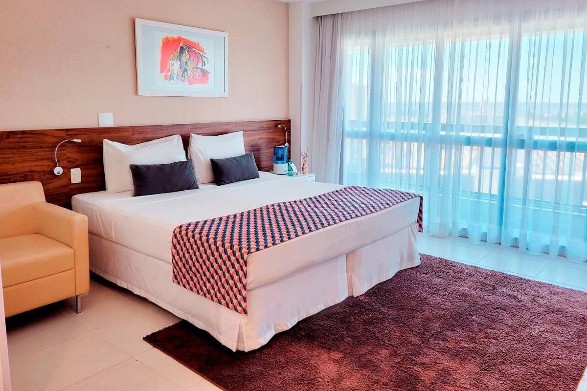 A hotel room with a bed, a chair, a bedside table, a lamp, a window with curtains, and a painting on the wall.