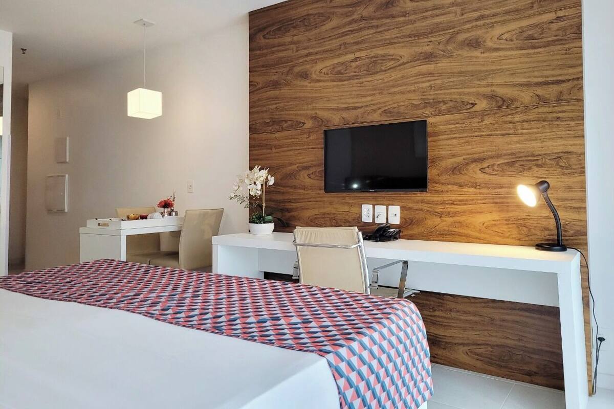 A hotel room with a bed, a desk, a chair, a TV mounted on a wooden wall, and a desk lamp.