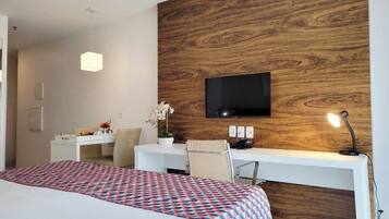 Quarto de hotel com cama, escrivaninha, cadeira, televisão presa a uma parede de madeira e abajur.