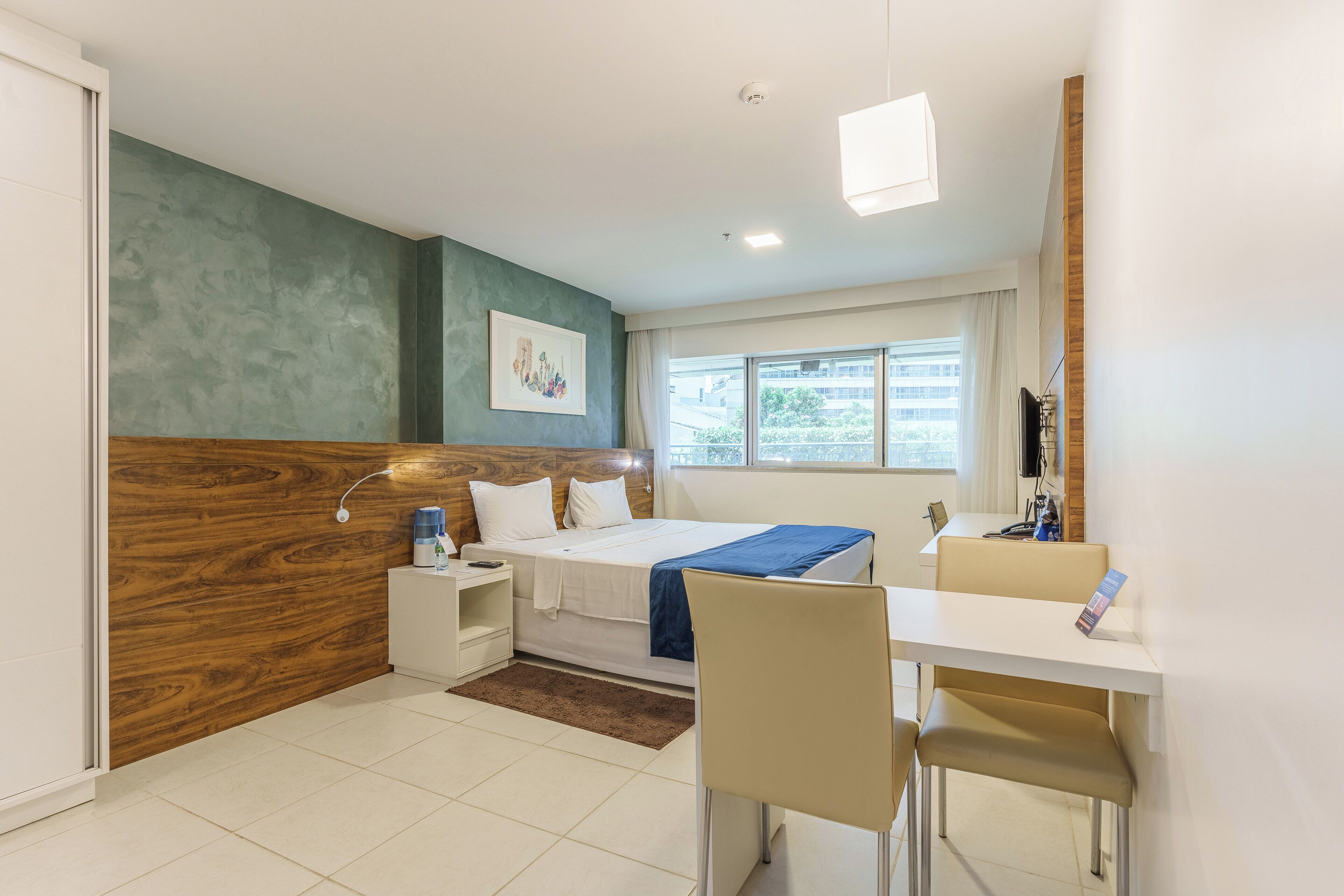 Quarto de hotel moderno com parede de destaque em madeira, cama com manta azul, escrivaninha com cadeira e janela com vista para edifícios.