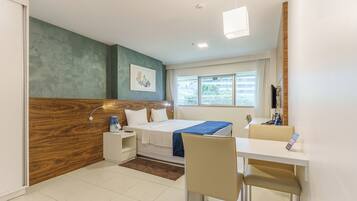 Quarto de hotel moderno com parede de destaque em madeira, cama com manta azul, escrivaninha com cadeira e janela com vista para edifícios.