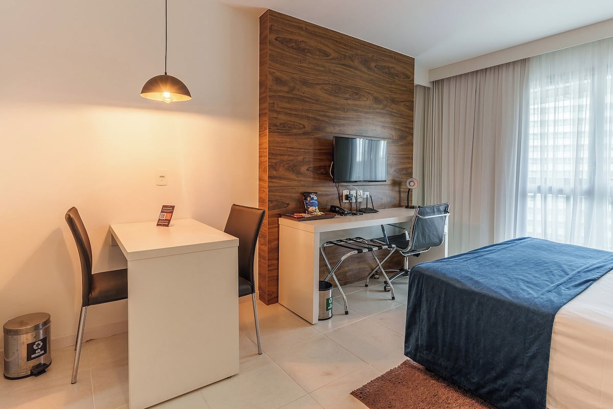A hotel room with a desk, chair, bed, TV, and a view of the city through the window.