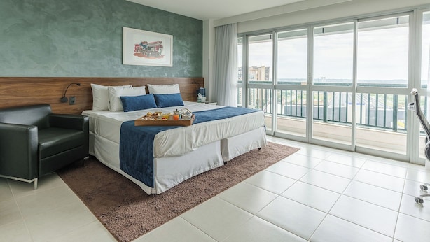 A modern hotel room with a large bed, a bedside table, a chair, and a view of the city through large windows. - Hplus Vision Executive (Brasília)
