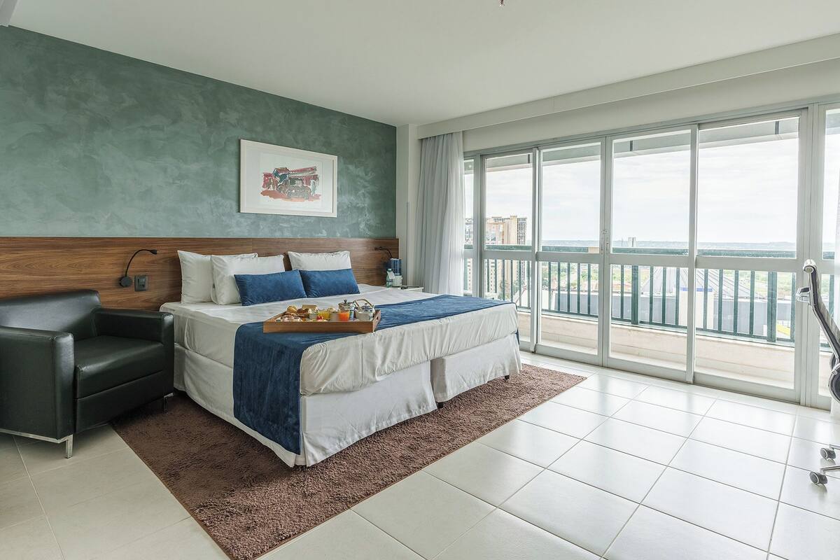 A modern hotel room with a large bed, a bedside table, a chair, and a view of the city through large windows.