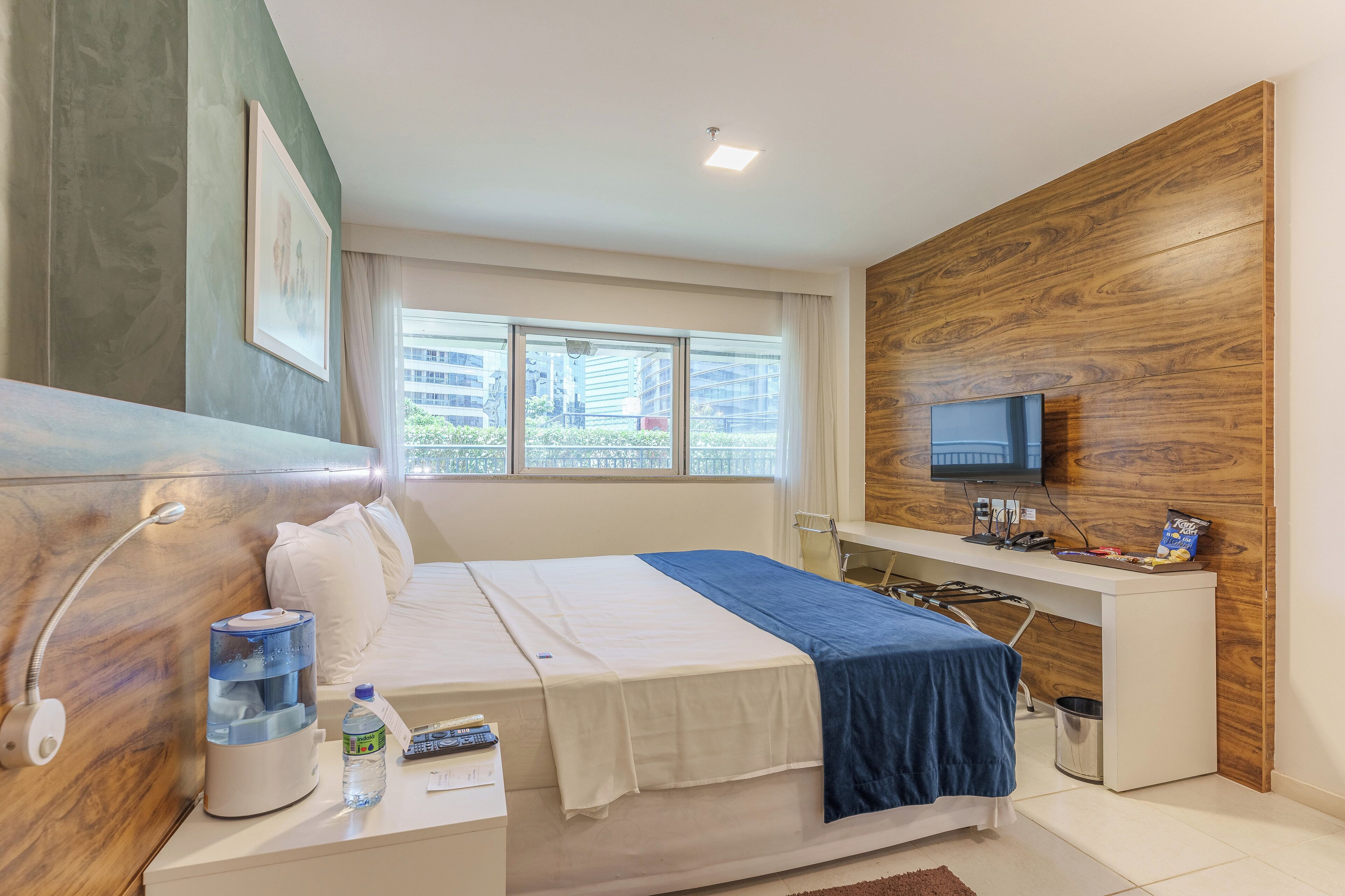 A modern hotel room with a bed, bedside table, water dispenser, and a TV mounted on the wall.