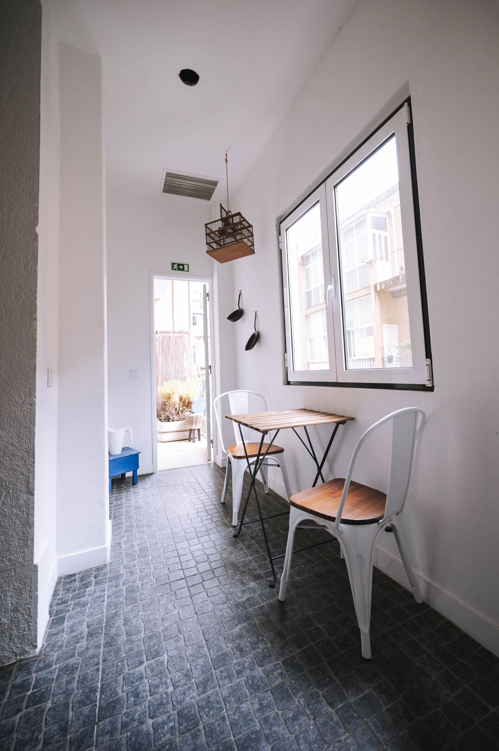 Un pasillo estrecho con suelo de baldosas, una mesa con sillas, un banco azul y una ventana con vistas a edificios.