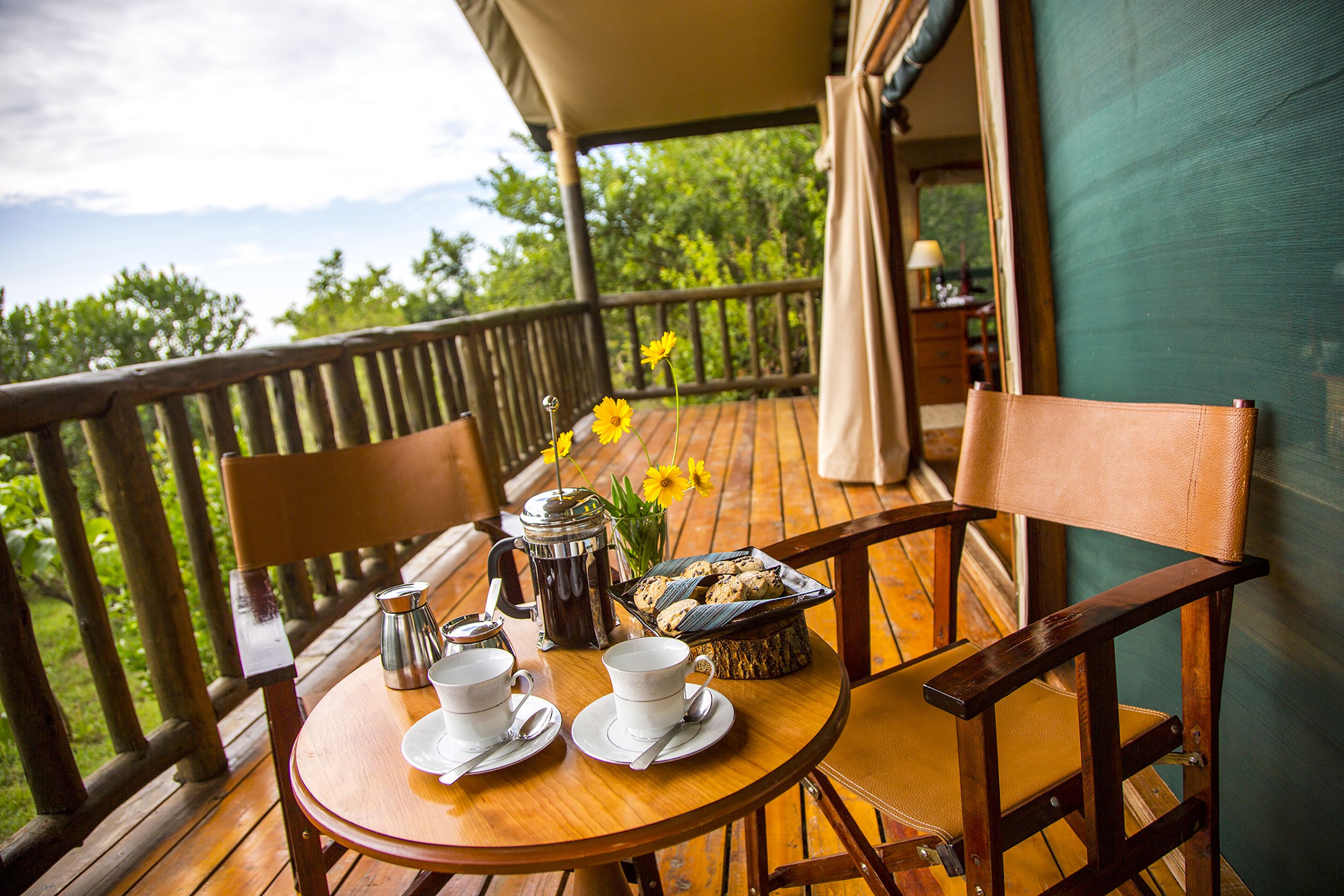 A wooden table with a coffee pot, cups, and a basket of cookies on a balcony with a view of trees.