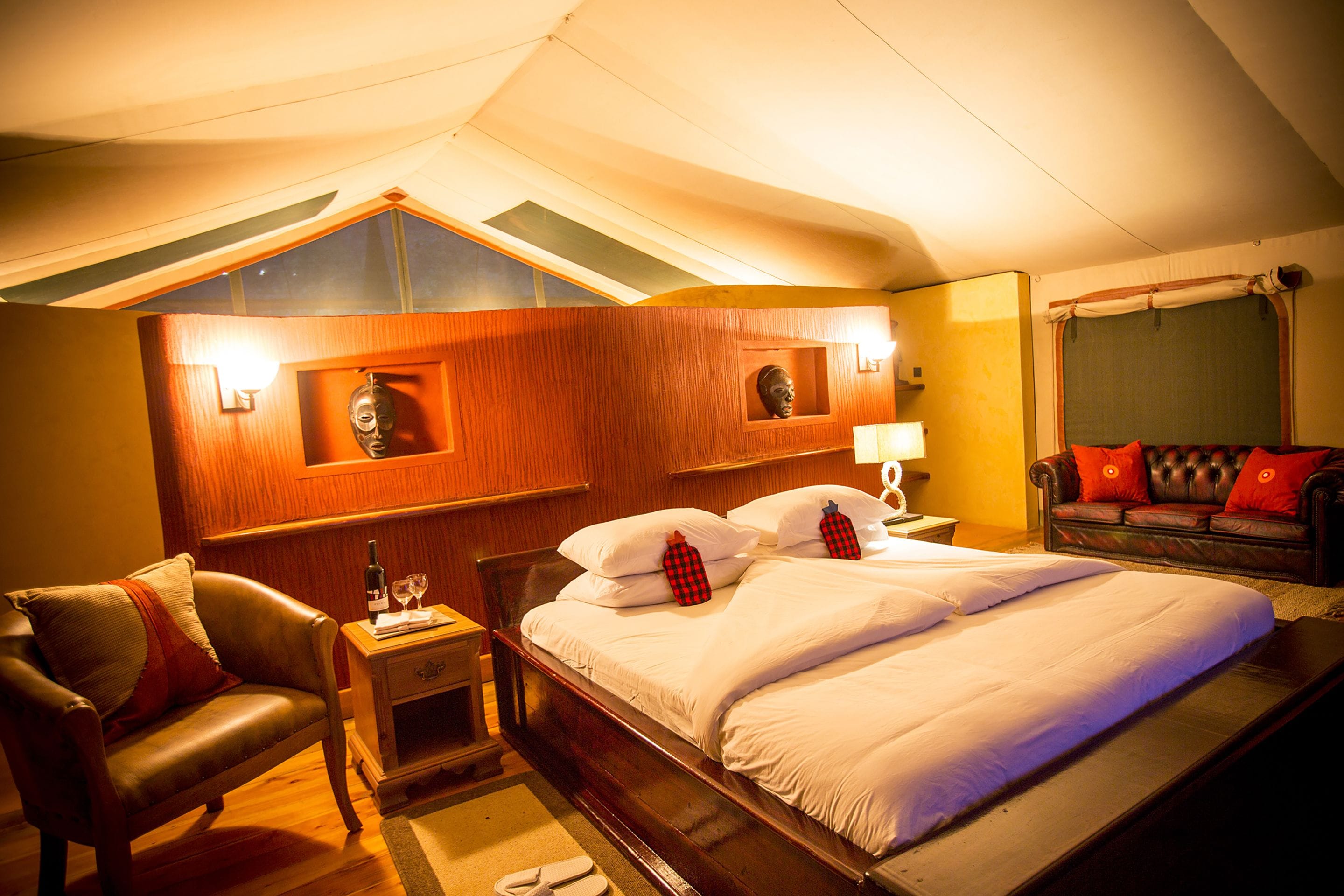 A hotel room with a large bed, a wooden headboard, a leather sofa, and a nightstand with a bottle and glasses.