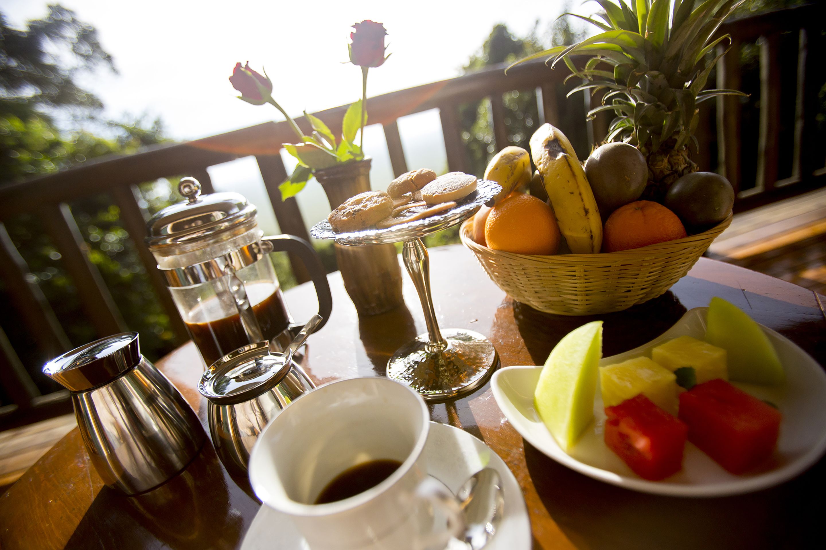 A table with a coffee pot, a cup of coffee, a glass of iced coffee, a plate of fruit, and a basket of bread and fruit.