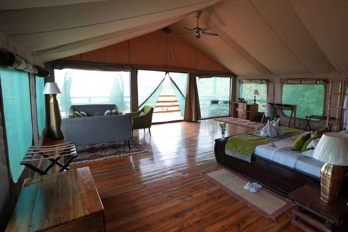 A spacious tented accommodation with a wooden floor, a bed with pillows, a sofa, a chair, and a desk.