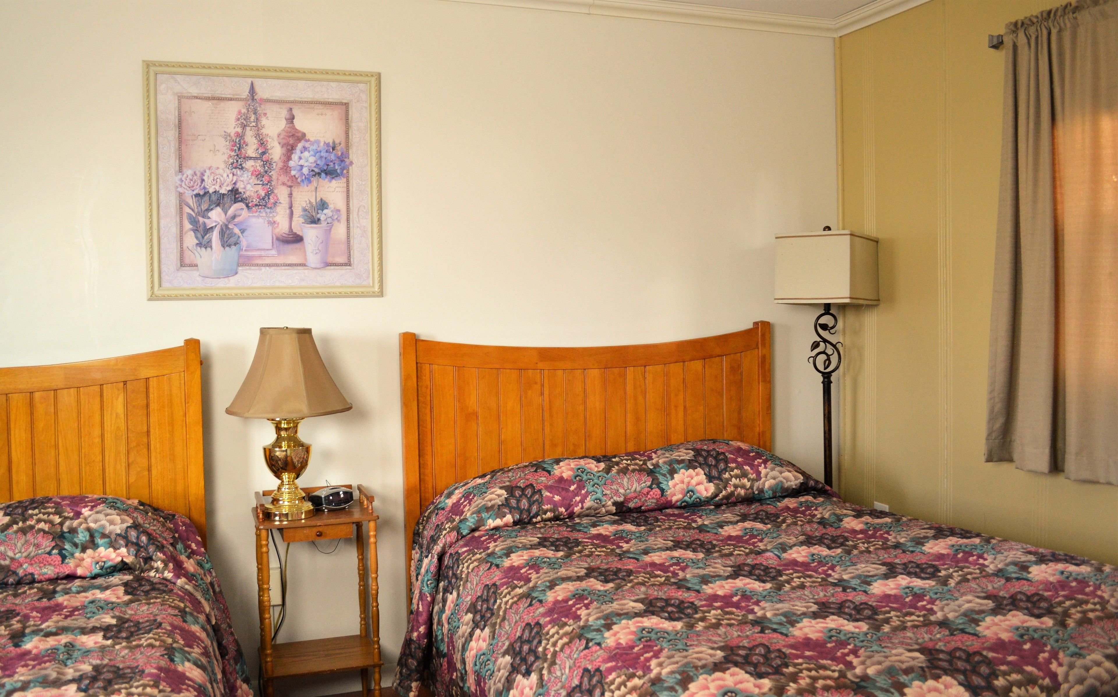 A bedroom with two wooden beds, a bedside table with a lamp, a framed floral artwork on the wall, and a floor lamp.
