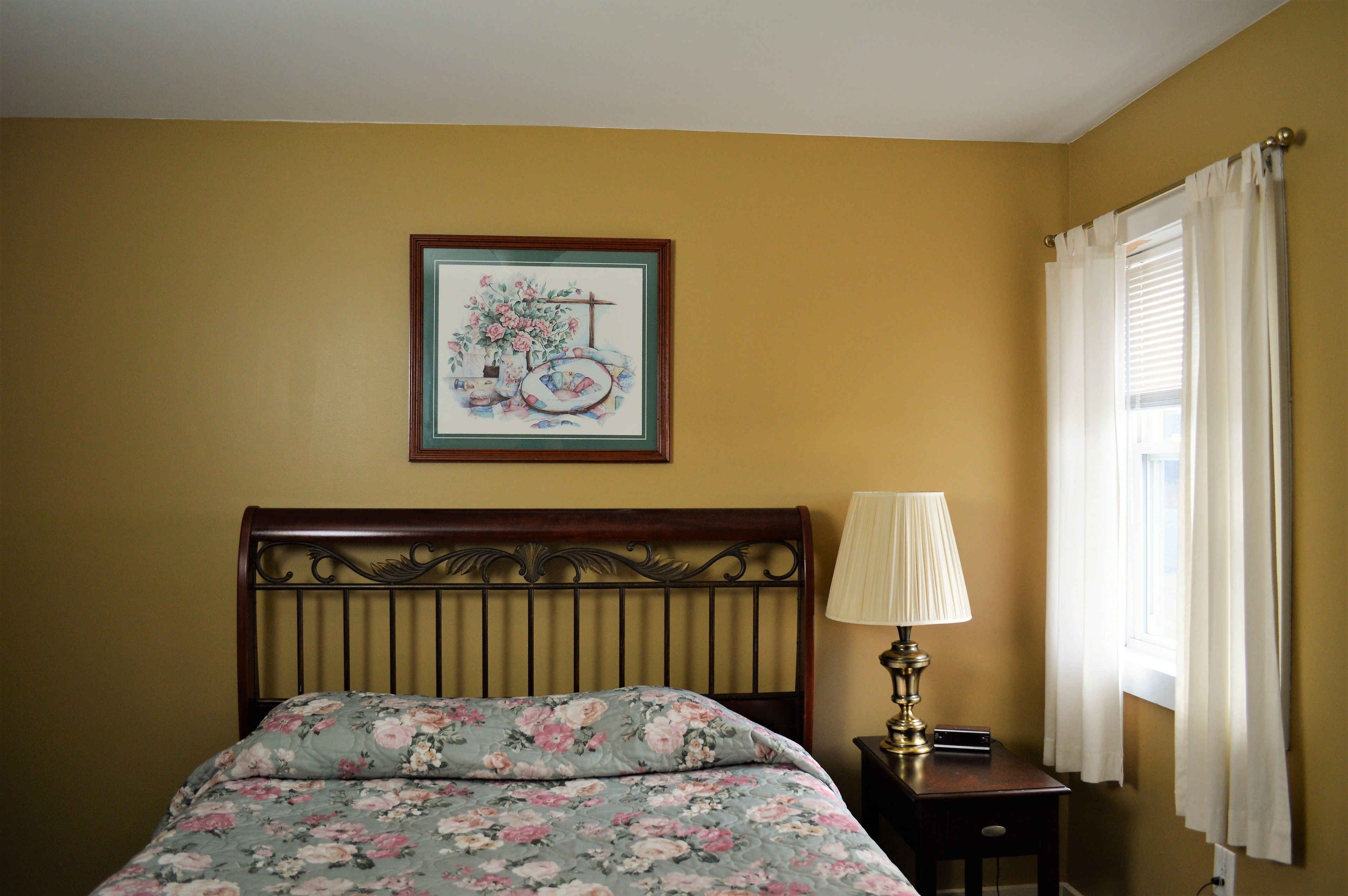 A bedroom with a floral bedspread, a framed picture on the wall, a bedside lamp, and a window with curtains.
