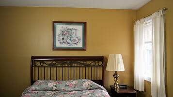 A bedroom with a floral bedspread, a framed picture on the wall, a bedside lamp, and a window with curtains.