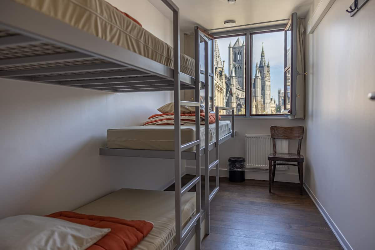 Quarto tipo dormitório com beliches, vista para um edifício histórico, radiador e uma cadeira.
