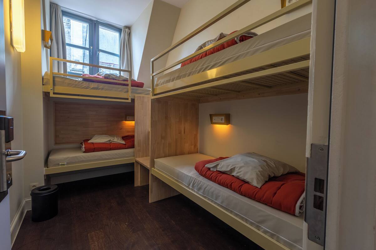 Um dormitório com beliches, uma janela com cortinas, uma porta e uma lixeira.