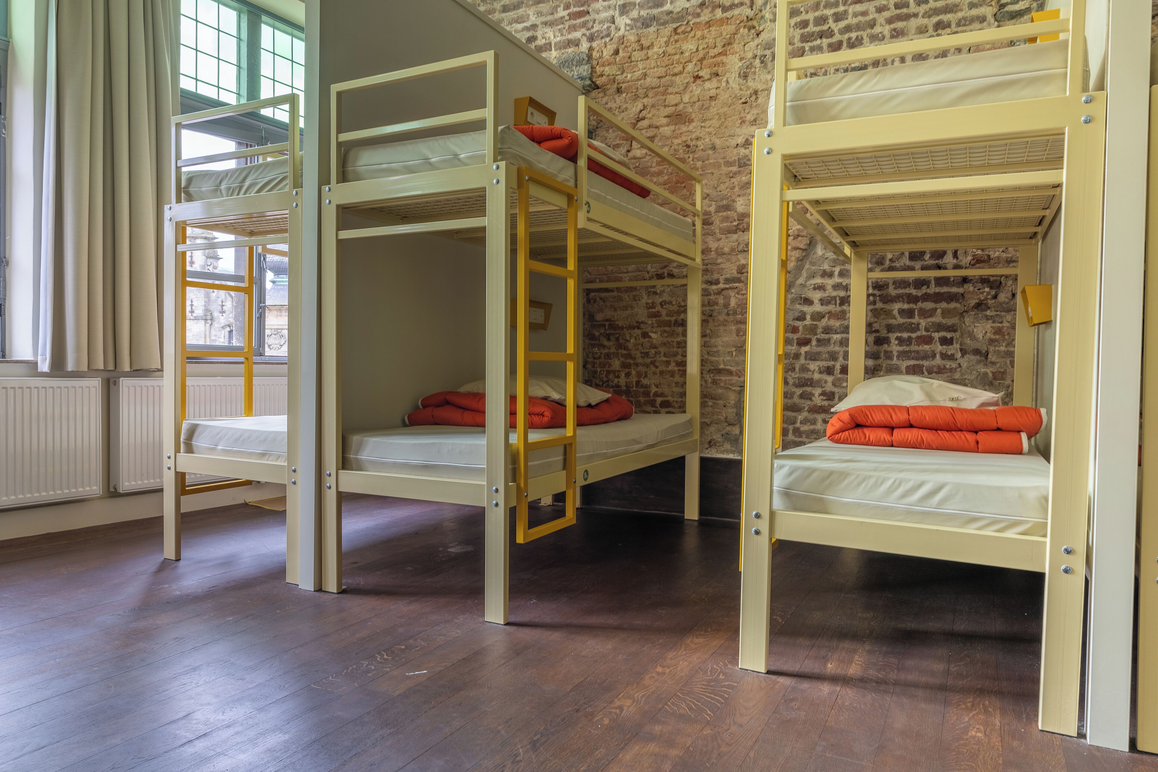 Une pièce avec trois lits superposés, un plancher en bois, un mur en briques et une fenêtre avec des rideaux.