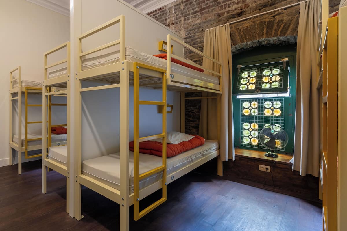 Um quarto com beliches, uma janela e um ventilador, e uma parede de tijolos.