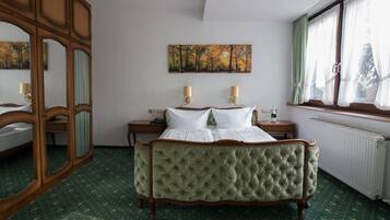 A room with a green upholstered bed, a wooden wardrobe, a painting of autumn trees, and a window with curtains.