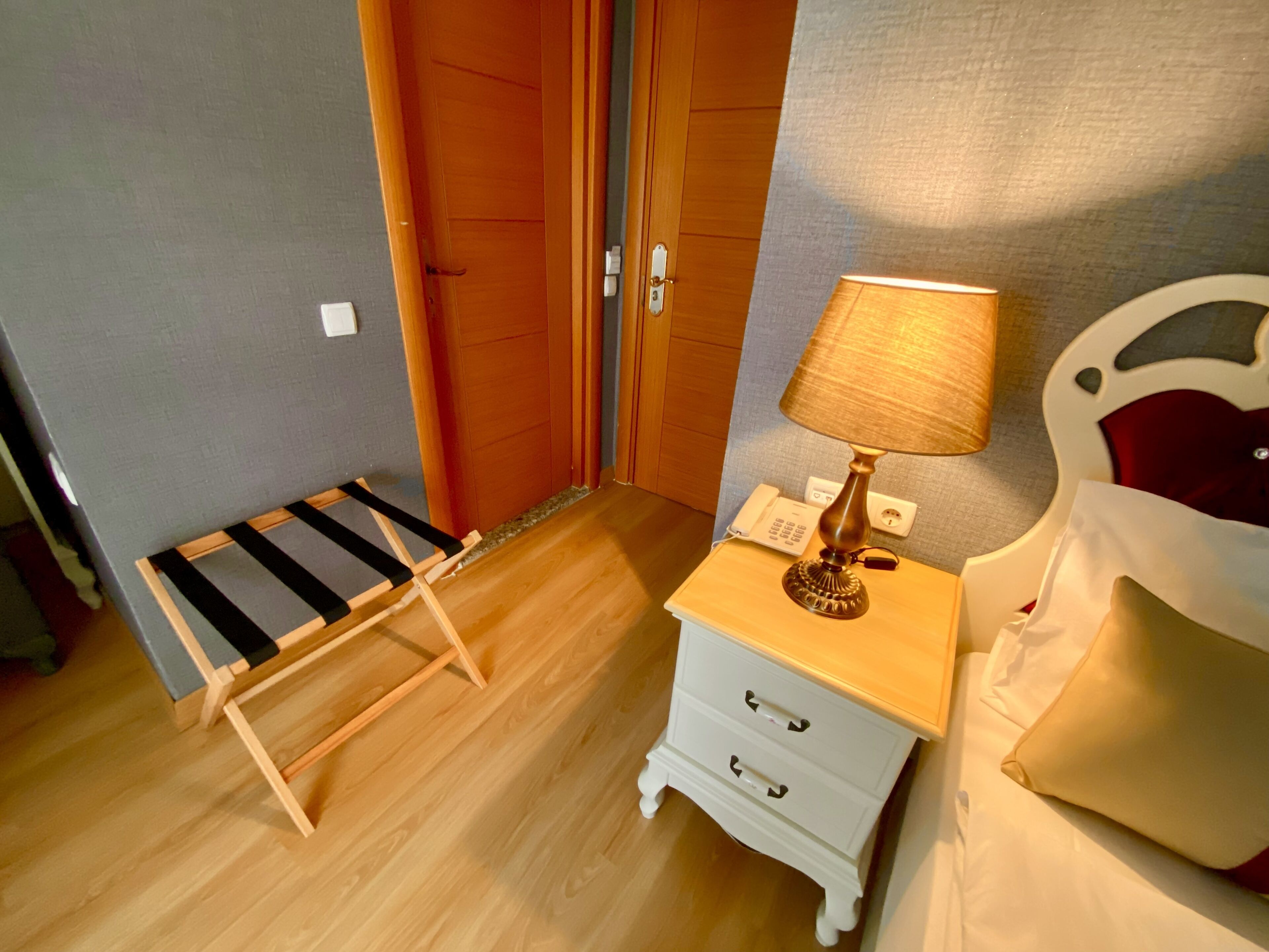 A room with a wooden floor, a bed with a headboard, a bedside table with a lamp, a small white dresser, and a striped bench.