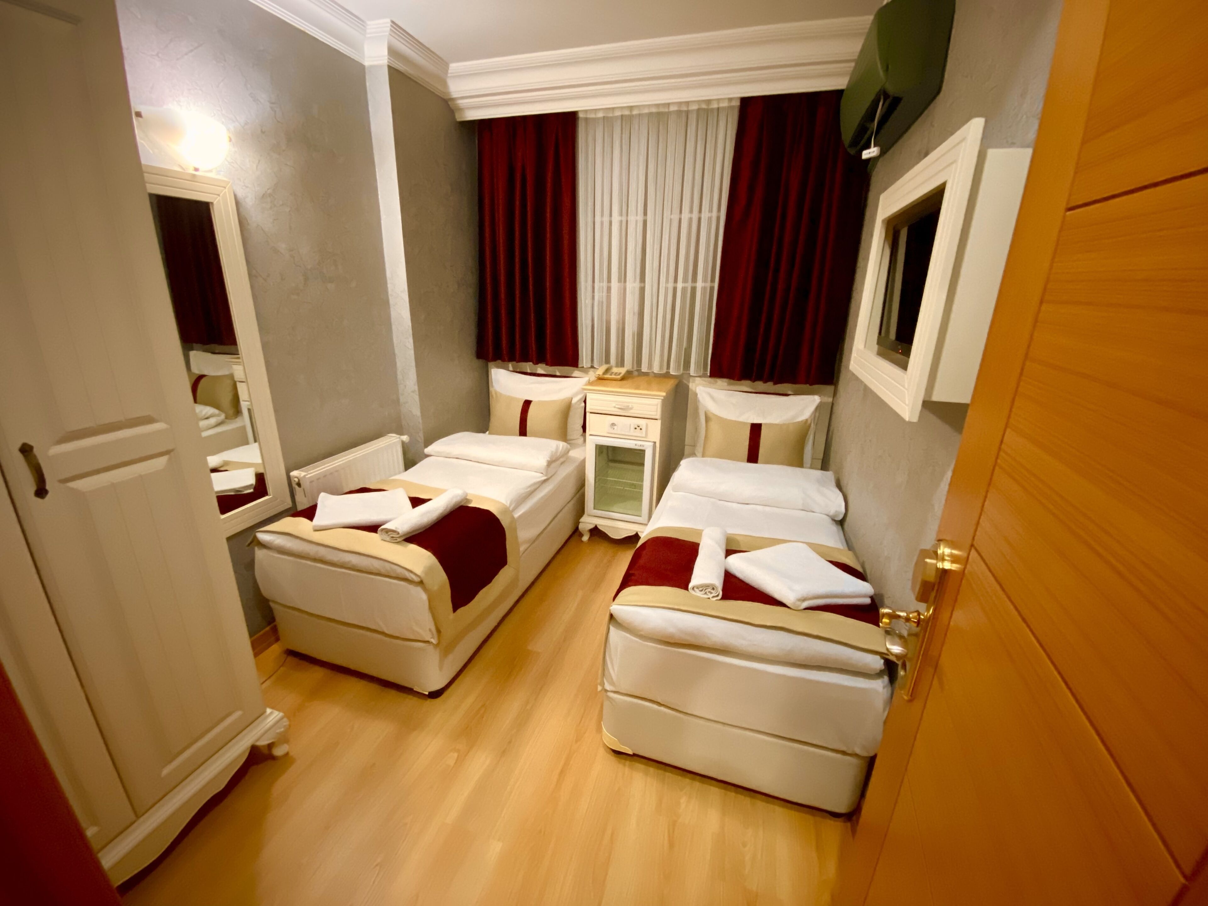 A compact hotel room with two single beds, a small bedside table, a nightstand, and a window with curtains.