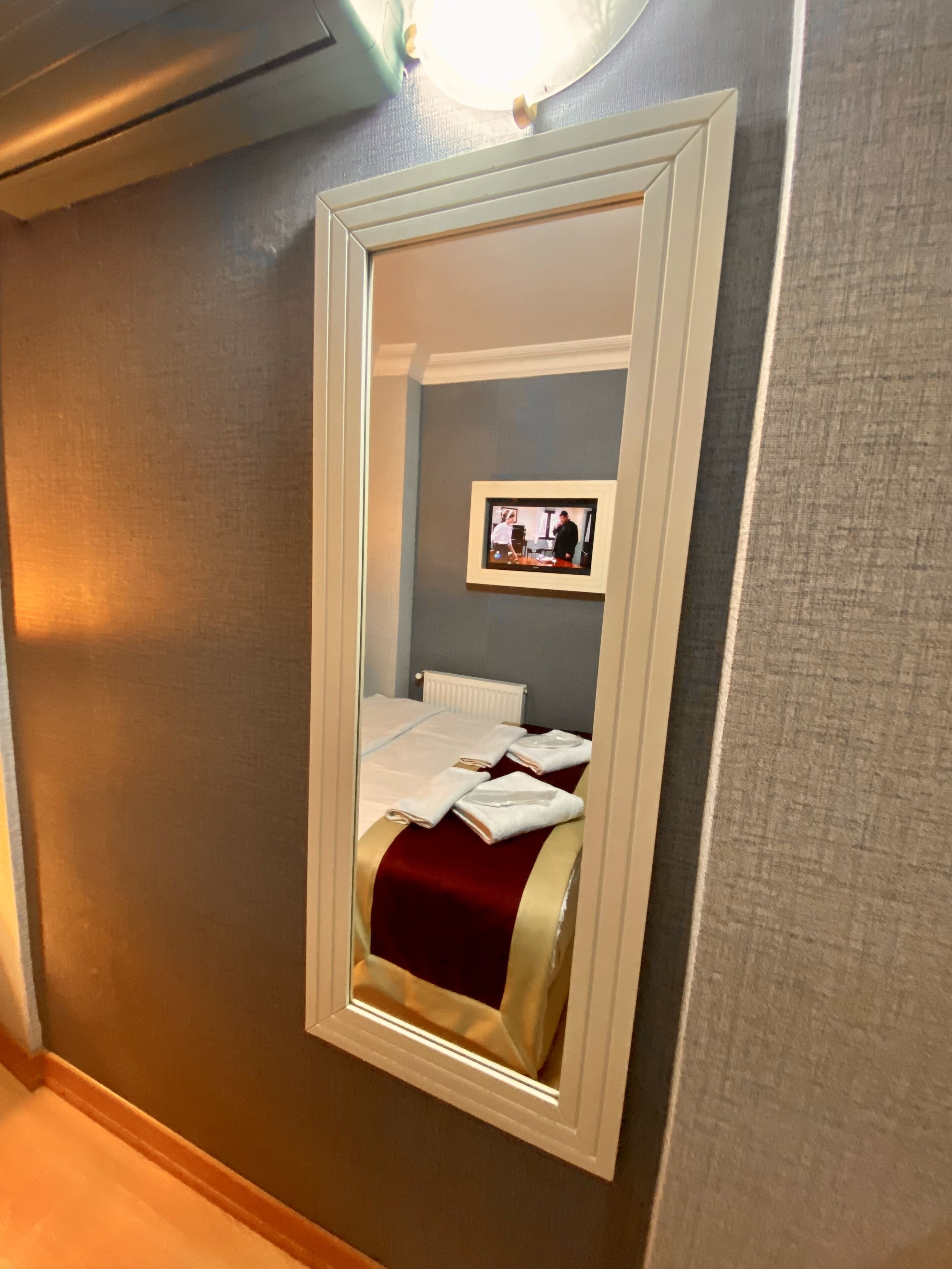 Een hotelkamer met een grote spiegel, waarin een bed met witte lakens en een rode dekbedovertrek wordt weerspiegeld.
