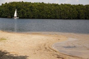 A sandy beach with a sailboat on the water and a forested shoreline in the background. - Hotel Villa Will (Isernhagen)