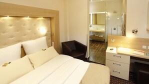 A hotel room with a large bed, a desk, a chair, and a bathroom visible through a glass door. - Hotel Villa Will (Isernhagen)