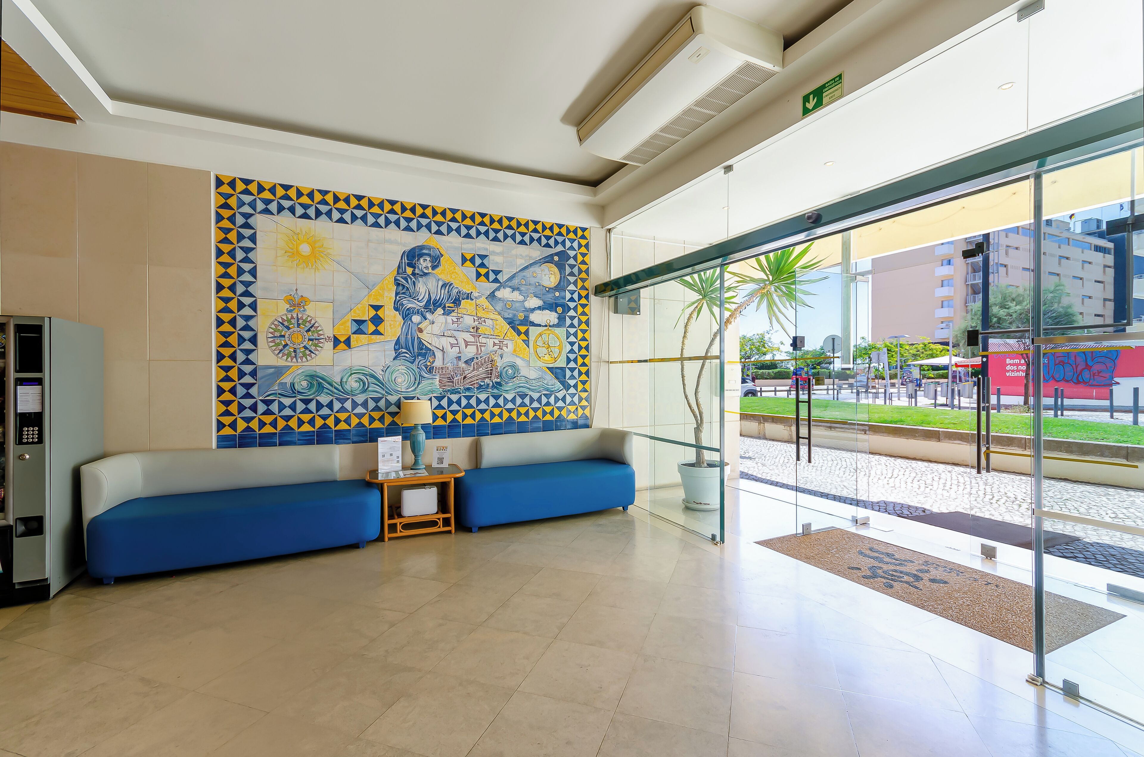 A hotel lobby with a large decorative wall, a blue sofa, a small table with a lamp, and a glass entrance leading outside.