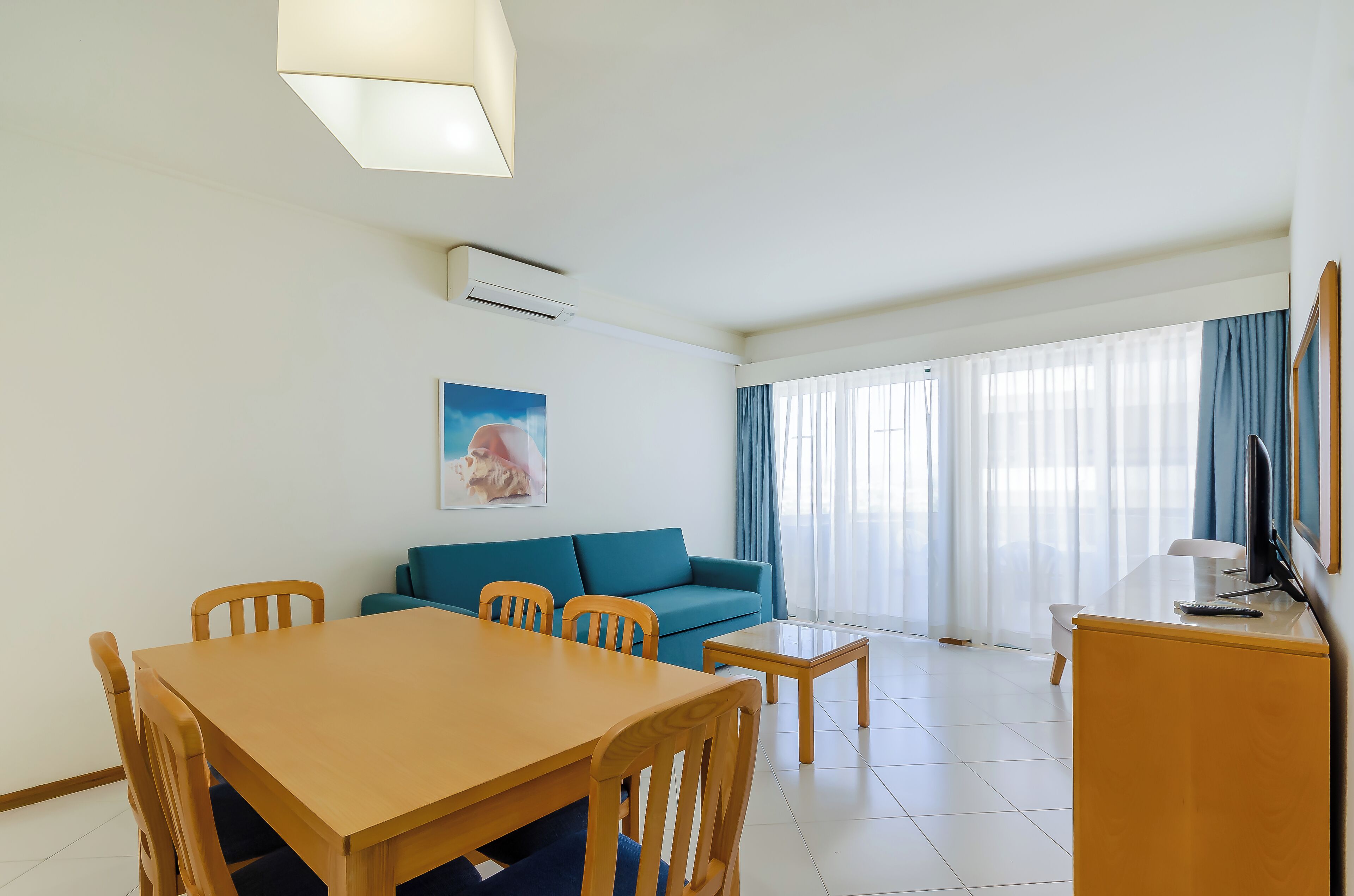 Ein Hotelzimmer mit Esstisch, Stühlen, einer Sitzecke, einem Fernseher und einem großen Fenster mit Vorhängen.
