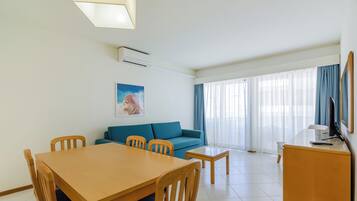 Ein Hotelzimmer mit Esstisch, Stühlen, einer Sitzecke, einem Fernseher und einem großen Fenster mit Vorhängen.