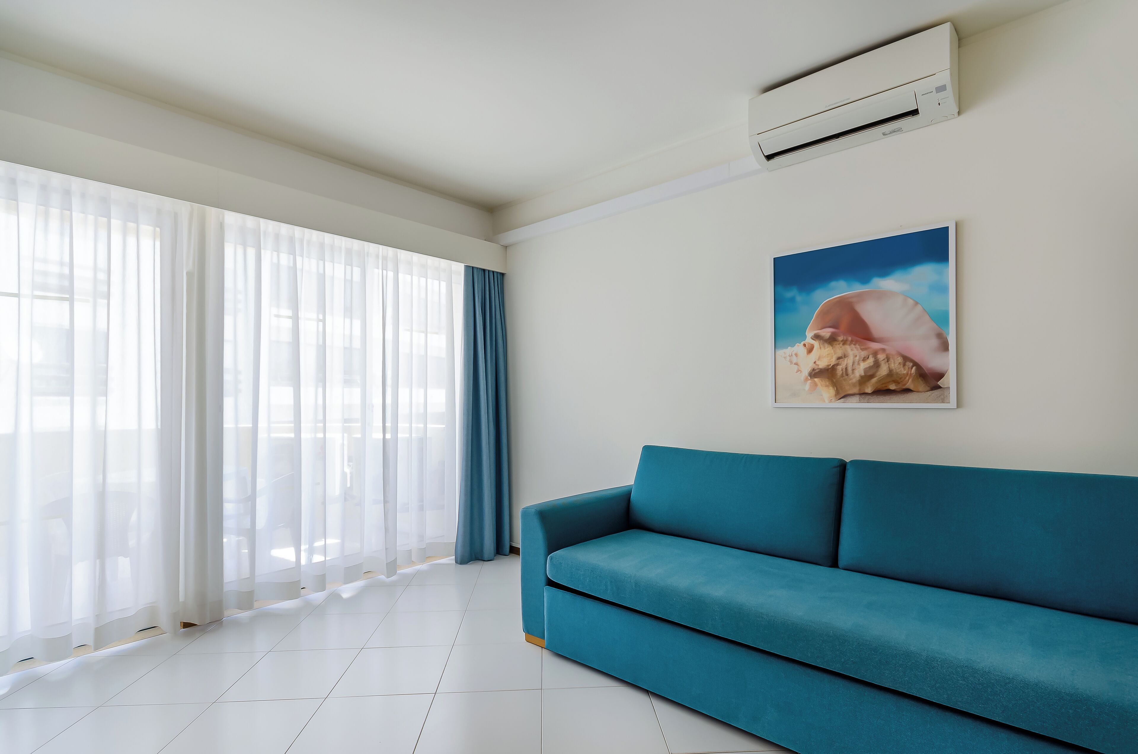 A modern living room with a teal sofa, large windows with sheer curtains, a wall-mounted air conditioner, and a framed picture of a shell.