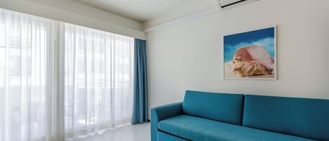 A modern living room with a teal sofa, large windows with sheer curtains, a wall-mounted air conditioner, and a framed picture of a shell.