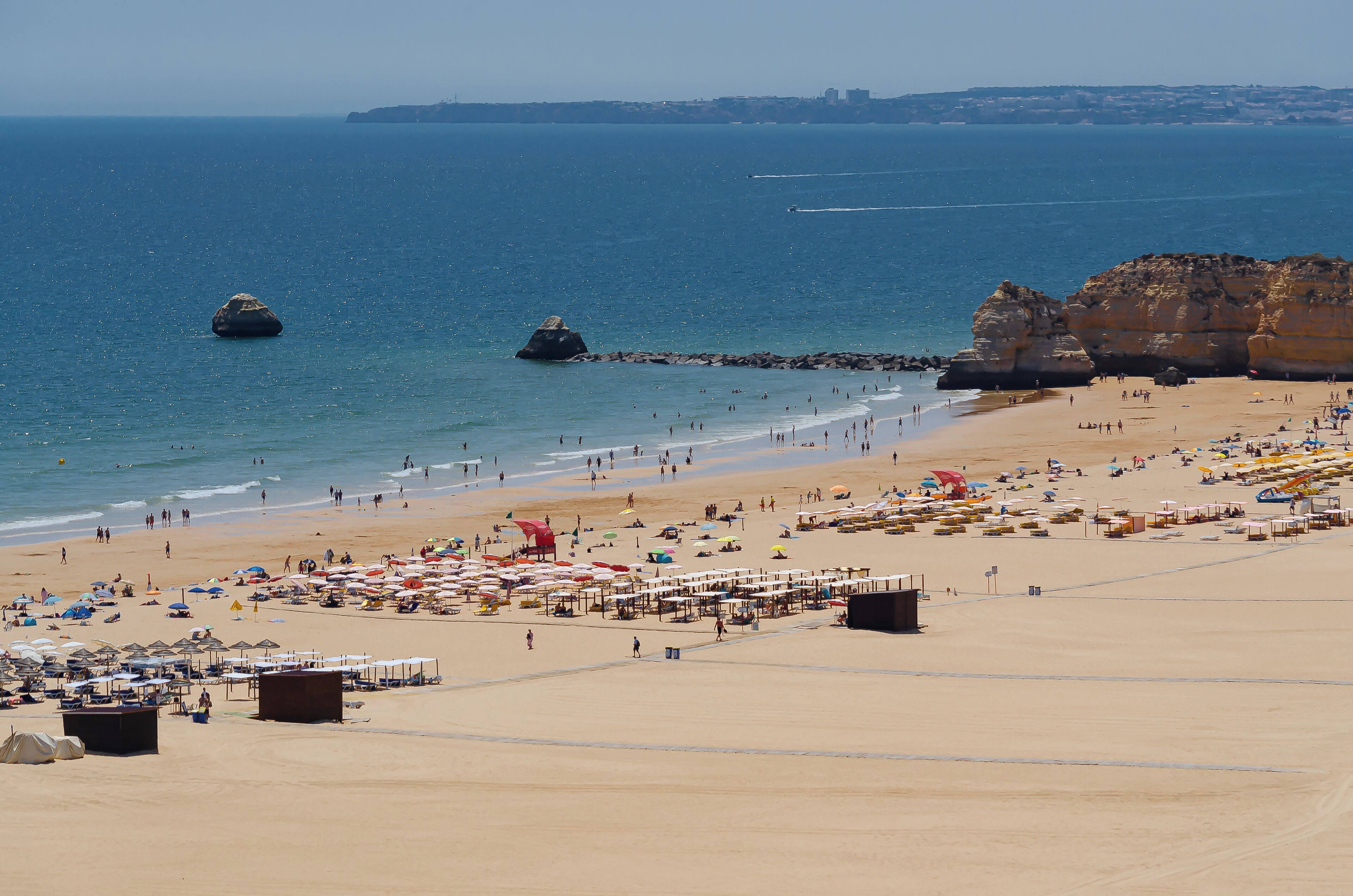 A beach with clear blue water, rocky formations, and numerous umbrellas and beachgoers.