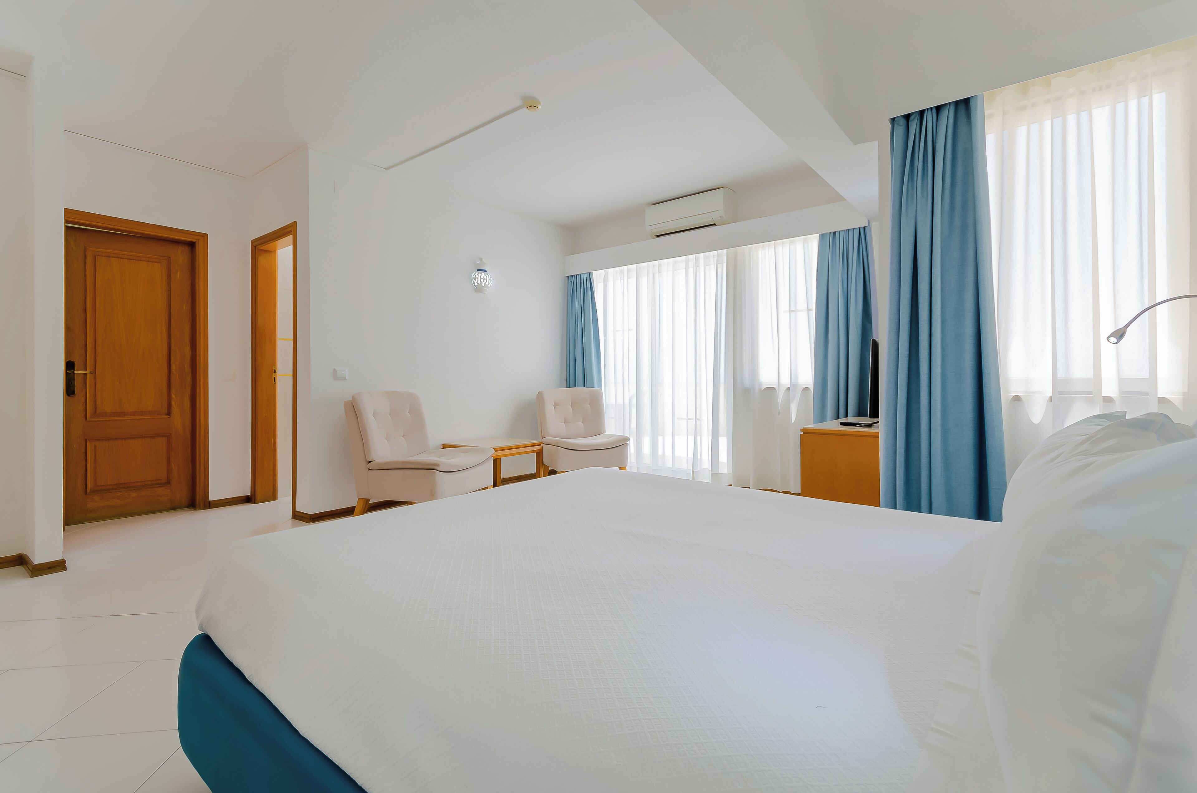 Une chambre d’hôtel avec un grand lit, deux chaises, un bureau et une fenêtre avec des rideaux.