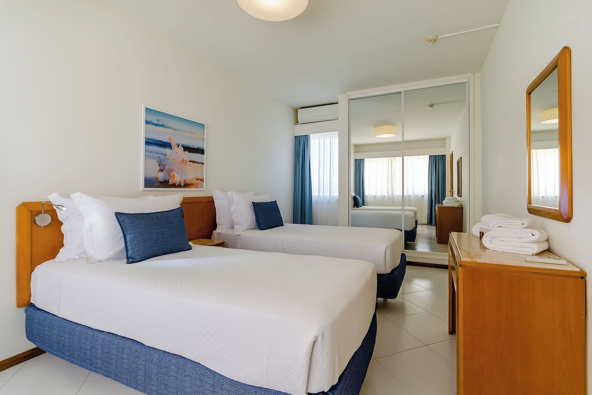A hotel room with two beds, a wooden headboard, a bedside table with towels, a large mirror, and a painting of a beach scene.