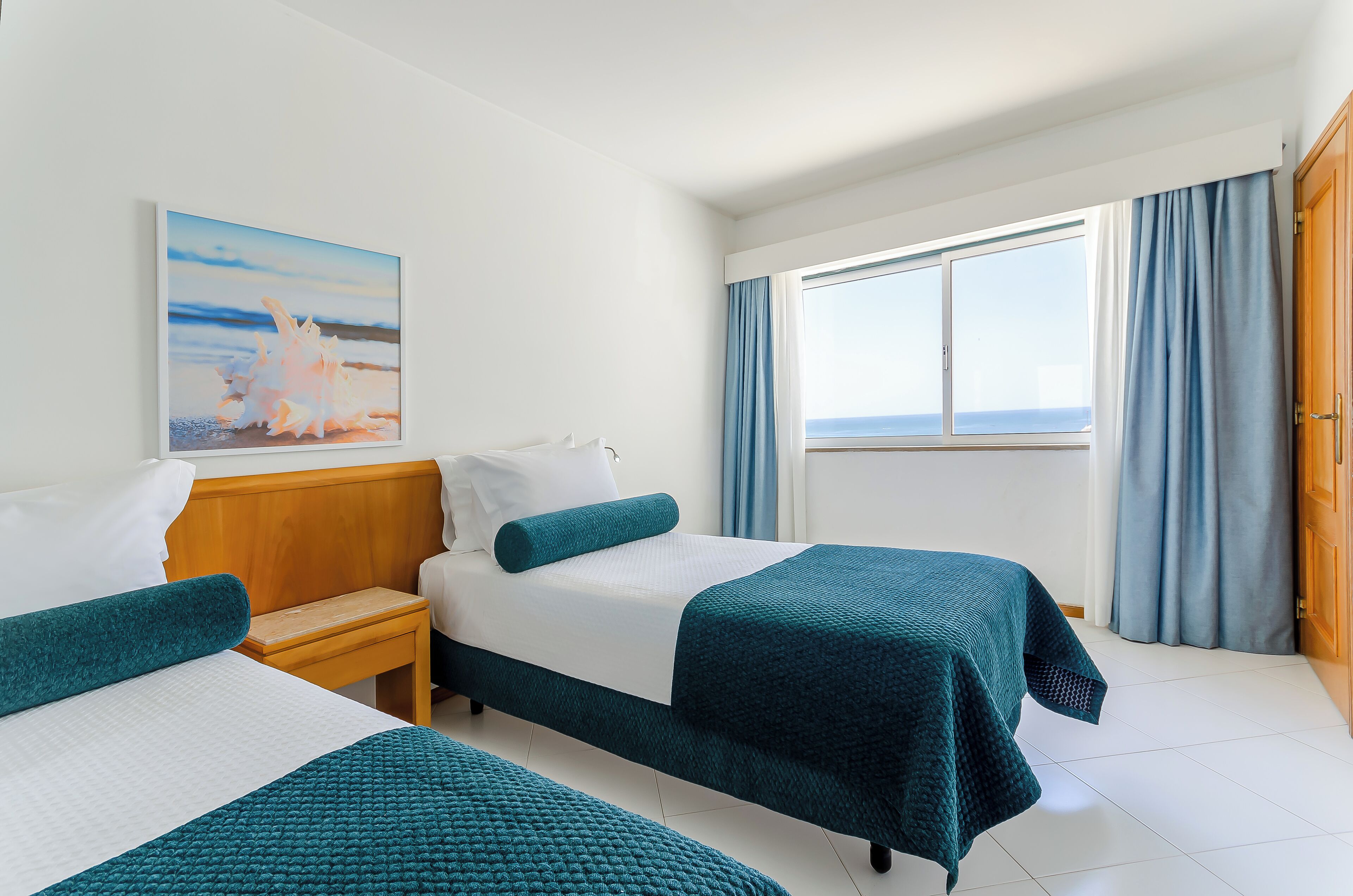 Habitación de hotel con dos camas, cabecero de madera, un cuadro de una playa, una ventana con cortinas y vista al mar.