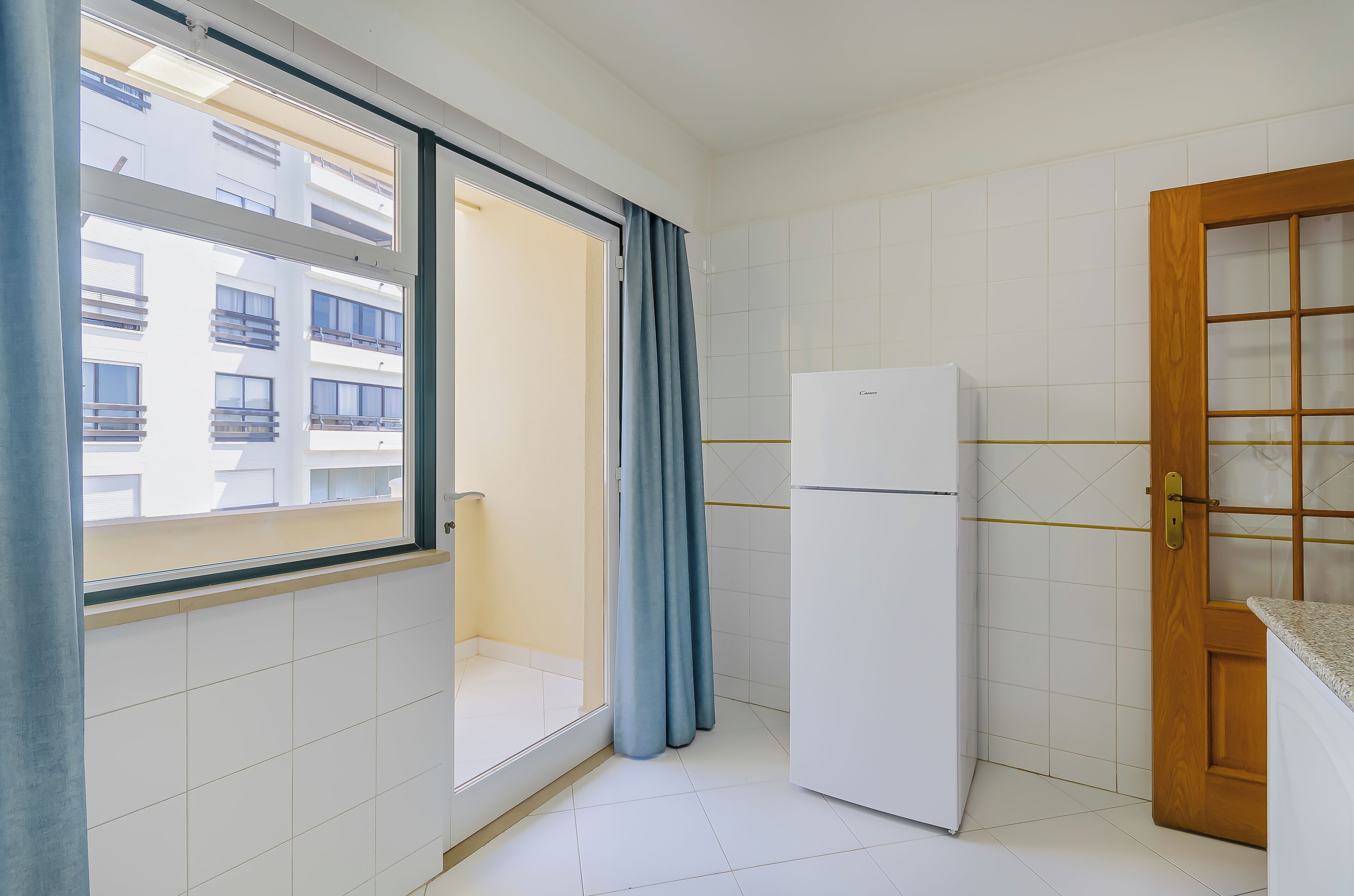 Eine kleine Küche mit Kühlschrank, einem Fenster mit Blick auf ein Gebäude und einer Holztür.