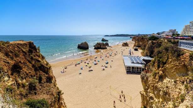 A beach with a sandy area, rocky cliffs, and a building with a roof. - TURIM Algarve Mor Hotel (Portimão)