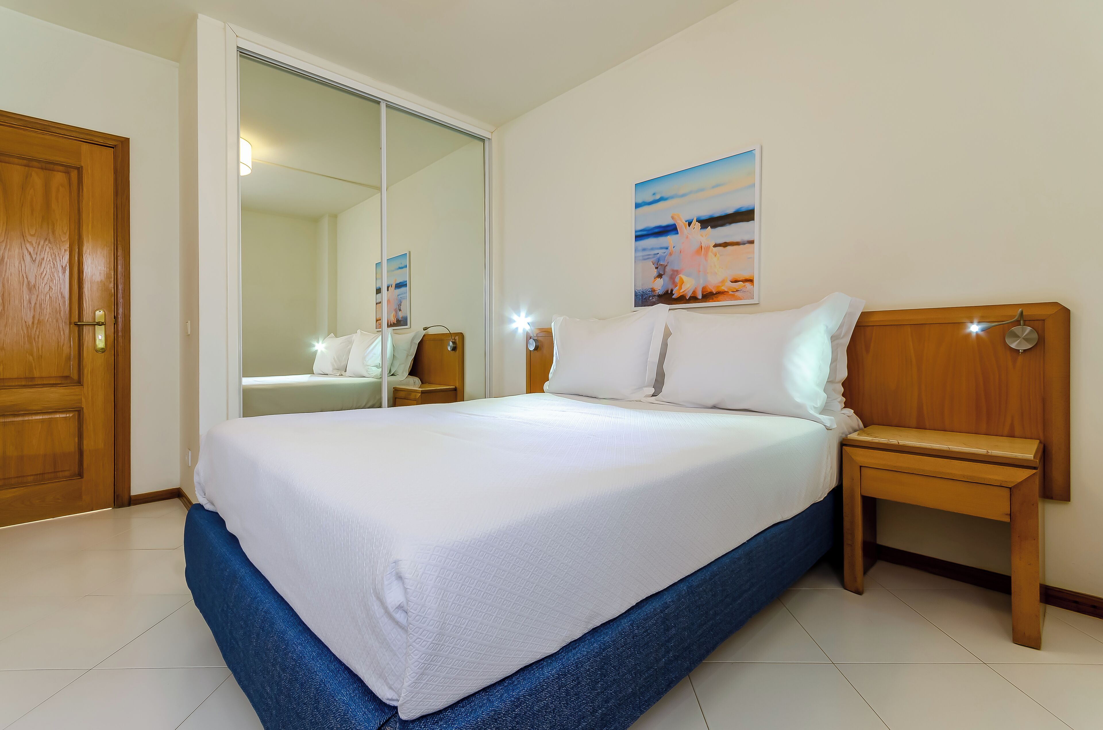 A hotel room with a large bed, a wooden headboard, a nightstand, a bedside table, a mirror, and a painting on the wall.
