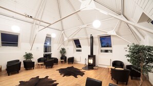 Amplia habitación con techo alto, suelo de madera y mobiliario de cuero negro. Tiene una chimenea, una planta en maceta y una alfombra grande.