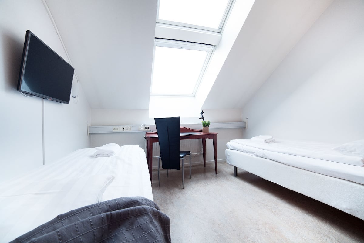 A small, well-lit room with two beds, a desk, and a chair.