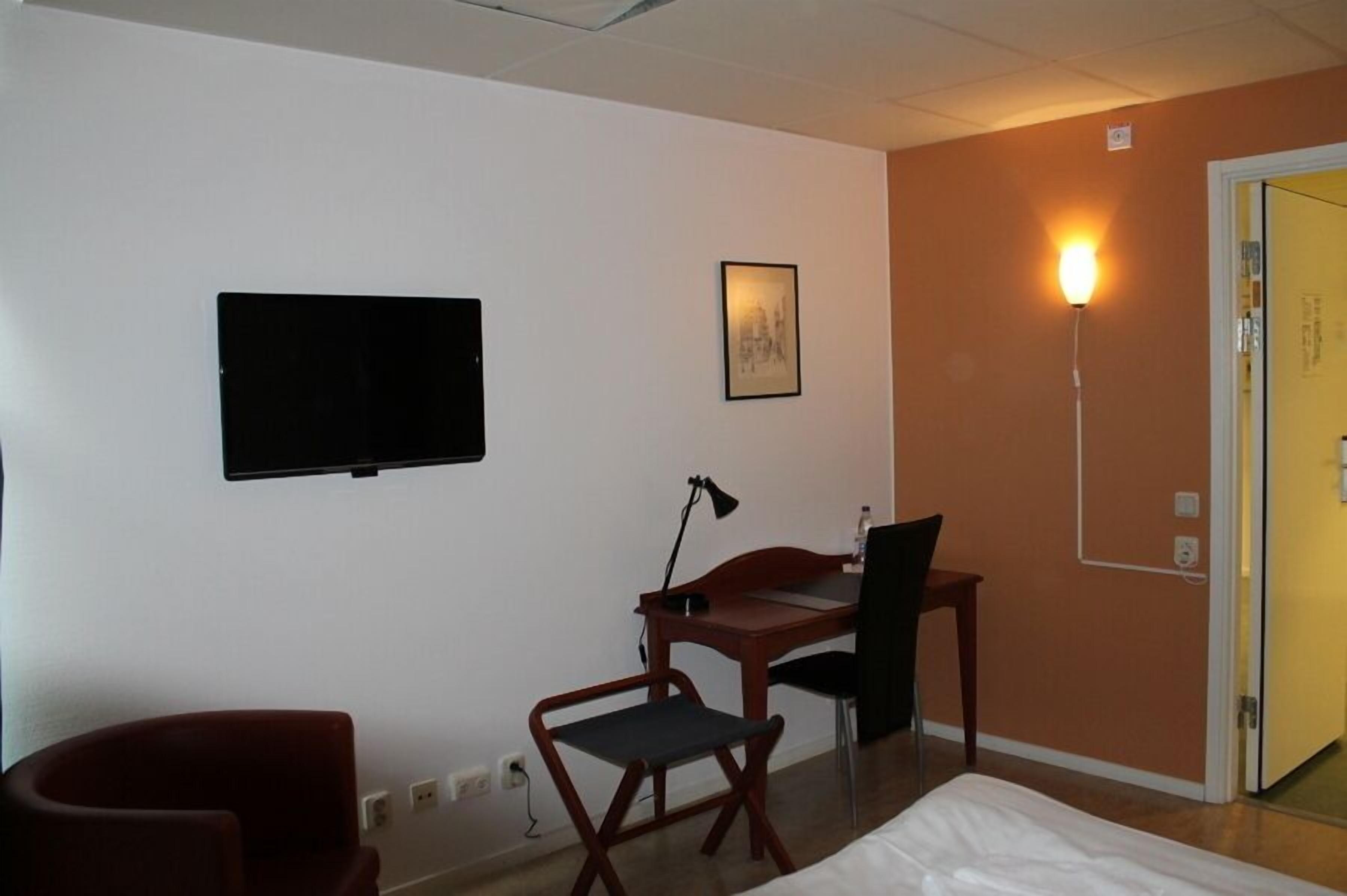 A hotel room with a bed, a desk with a chair, a television mounted on the wall, and a lamp.